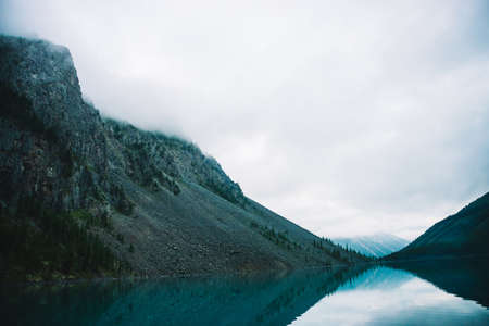 Giant cloud above rocky mountainside with trees in fog. Amazing mountain lake. Mountain range under cloudy sky. Wonderful rocks in mist. Morning landscape of highland nature. Low clouds in mountains.の写真素材