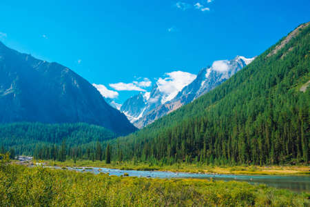 Mountain creek in valley before beautiful glacier. Snowy rocks behind mountains with conifer forest. Huge clouds on giant snowy mountain top under blue sky. Atmospheric landscape of highland nature.の写真素材
