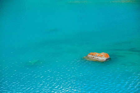 Orange mossy stone in azure mountain lake. Textured background of clean shiny water surface in...