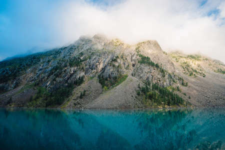 Giant cloud above rocky ridge with trees in sunlight. Amazing mountain lake. Mountain range under...