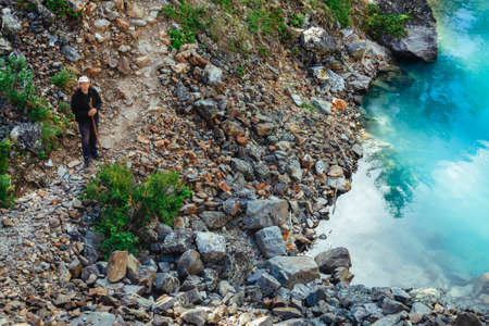 Alone traveler with wooden stick on pathway near clear azure mountain lake. Hiking in mountains....