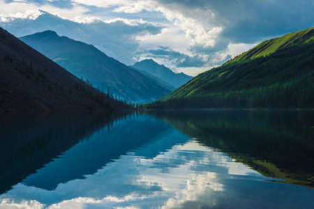 Ghostly mountain lake in highlands at early morning. Beautiful misty silhouettes of mountains and clouds reflected in clear water surface. Smoke of campfires. Amazing landscape of majestic nature.の写真素材