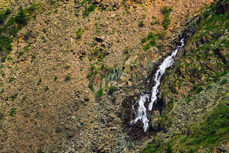 Detailed natural texture of slope from loose stones. Mountain water stream flows down mountainside. Small waterfall on rock with vegetation. Unusual landscape of majestic nature.の写真素材