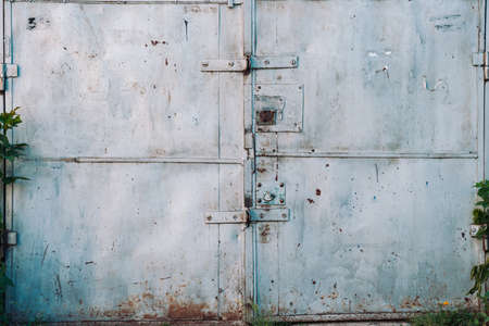 Closed imperfect rust metallic garage gate close-up. Damage texture of locked rusty iron door. Grungy metal surface. Textured background of rough faded uneven steel gates. Obsolete metal surface.の素材 [FY310122912723]