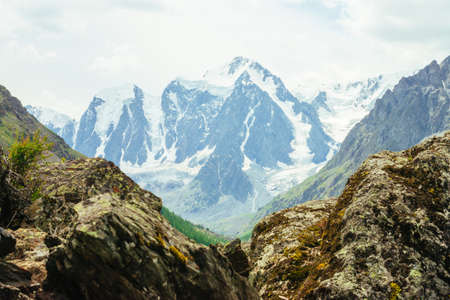 Wonderful glacier on rock background. Giant snowy mountain top behind rocky mountains under cloudy sky. Huge mountainside. Rich vegetation of highlands. Atmospheric landscape of majestic nature.の写真素材