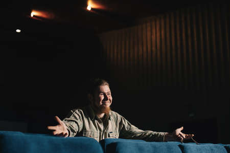 Director by one in dark theater hall. Portrait of creative man in low key. Alone art person in concert hall. Surprised critic at premiere. Happy brutal bearded man with glasses in culture house.の写真素材