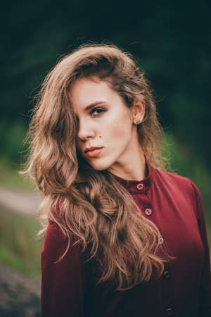 Beautiful girl with curly natural hair enjoy nature in forest. Dreamer lady in burgundy dress on green background. Inspired girl. Female face portrait. Sun in hair in autumn. Vintage portrait.の写真素材