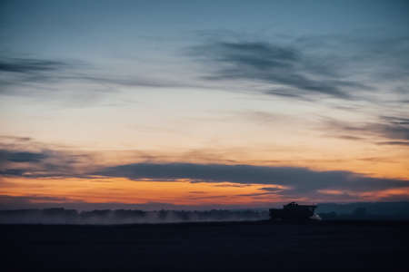Silhouette of harvester machine to harvest wheat on sunset. Combine harvester driving on field on sunrise. Beautiful dawn sky above wheat field. Combine working in dusk. Wonderful twilight landscape.の写真素材