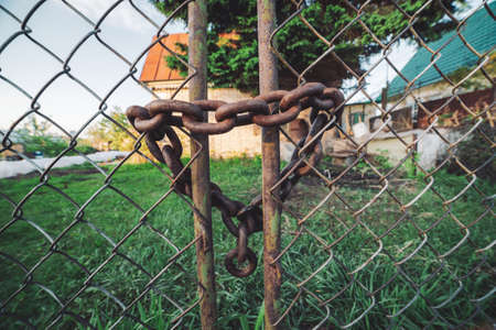 Iron rusty chain-link grid fence. Rust on metallic locked gate. Chain with lock close-up. Grungy chain in heart shape. Country house behind steel mesh fence. Rustic rough background with copy space.の写真素材