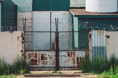 Picturesque industrial building close-up. Metallic rusty gate and concrete fence with barbed wire. Entrance in old factory. Closed area. Old industrial object is overgrown with grass. High metal doorsの写真素材