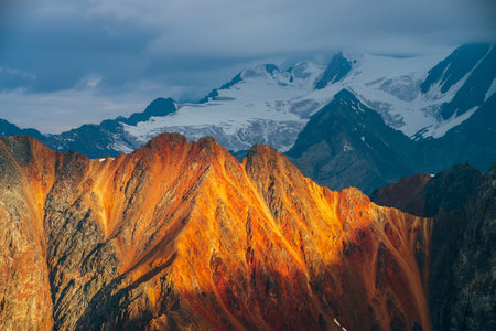 Atmospheric alpine landscape with red rockies in golden hour. Scenic view to big orange rocks and giant snowy mountains with glacier in sunrise. Wonderful highland scenery. Flying over mountains.の写真素材