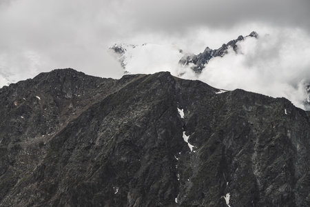 Atmospheric minimalist alpine landscape with big rocky mountain wall and snowbound peak in low...