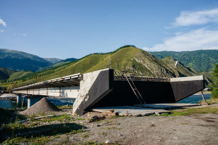 Big bridge construction over mountain river close-up. Vivid sunny landscape with unfinished bridge and big fast mountain river on background of beautiful green hills with forest under blue sky.の写真素材
