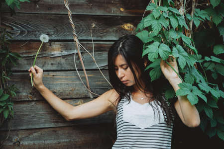 Lonely inspired girl with blowball flower near wooden wall with greenery. Country girl with...