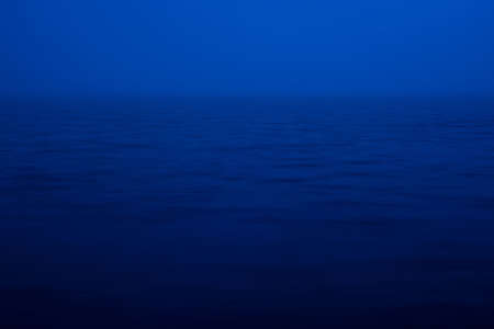 Natural texture of deep blue calm water in dusk close up. Night sea of blue classic color. Water ripple nature background. Meditative image of thick fog above lake. Soft light shines on water surface.の写真素材