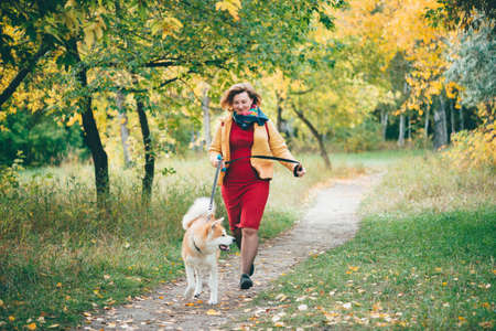 Girl with cute young foxy dog runs along pathway among fall foliage in autumn park. Running with ginger husky. Walking pet with owner in fresh air. Fat woman and white red husky dog in outdoors.の写真素材