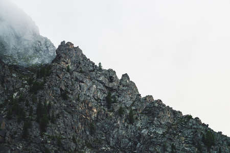 Ghostly atmospheric view to big cliff in cloudy sky. Low clouds among giant rocky mountains. Mysterious place at early foggy morning. Minimalist scenery with beautiful rockies. Dramatic bleak fog.の写真素材