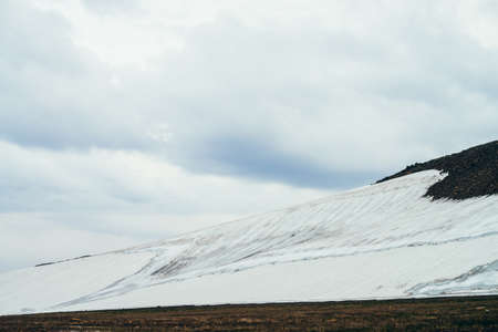 Atmospheric minimalist alpine landscape with wide glacier on rocky mountain slope under cloudy sky. Wonderful mountain scenery with snowy mountainside in highland valley. Rocky hills with snow.の写真素材