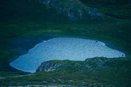 Small mountain lake in highland valley. Atmospheric minimalist mountain landscape with blue water among green mosses on rocks. Bleak atmosphere of dark valley. Beautiful ghostly place in mountains.の写真素材
