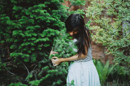 Gentle inspired girl caresses fir needles in garden in spring time. Young country girl with black long hair among summer greenery. Beautiful dreamy female portrait on vivid nature green background.の写真素材