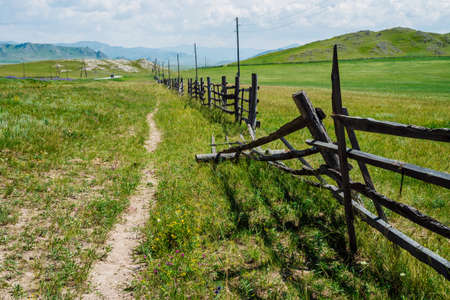 Broken wood fence and poles with wires along vast field in mountains in sunny day. Beautiful sunny alpine landscape with asphalt road along field behind long fence in highlands. Vivid mountain sceneryの写真素材