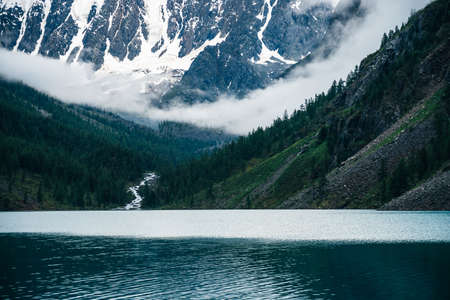 Beautiful big glacier, rocky snowy mountains among low clouds, coniferous forest on hills, mountain lake and highland creek. Atmospheric alpine scenery with high forest steep slopes and dense clouds.の写真素材