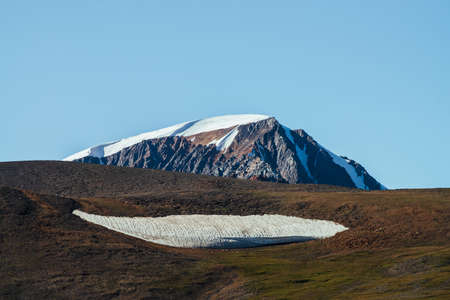 Giant snowy mountain behind green hill with big piece of firn. Great glacier under blue sky. Wonderful sunny highland scenery with huge mountain with snow. Beautiful minimalist alpine landscape.の写真素材