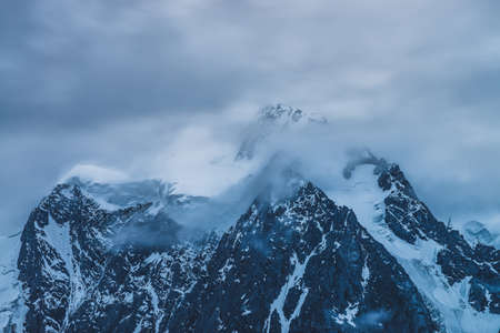 Mysterious dramatic alpine scenery with snowy mountain top inside low clouds in dusk. Bleak view to...