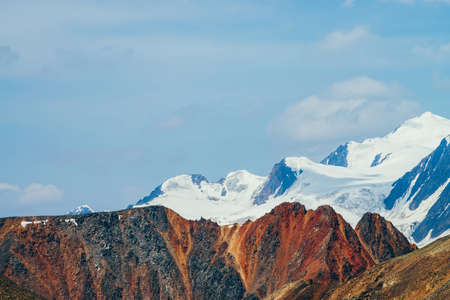 Minimalist beautiful alpine landscape with huge snowy mountain behind vivid pointed red craggy wall. Big glacier under blue sky. Atmospheric colorful highland scenery with giant red orange brown rock.の写真素材