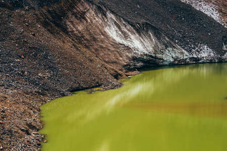 Stones on slope of glacier above mountain lake of acid green color. Beautiful emerald glacial lake and snowy slope of glacier with stones. Awesome alpine lake of unusual green tones. Amazing landscapeの写真素材