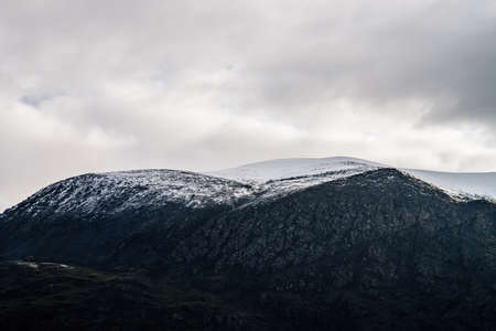 Great black mountain with white snow on top. Black and white dramatic landscape with snowy mountain under cloudy gray sky. Atmospheric alpine scenery with snow on rocky mountain in overcast weather.の写真素材