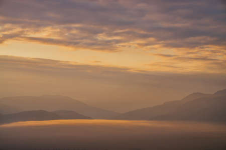 Scenic dawn mountain landscape with golden low clouds in valley among mountains silhouettes under cloudy sky. Vivid sunset or sunrise scenery with low clouds in mountain valley in illuminating color.の写真素材