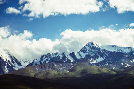 Awesome mountains landscape with sunlit high snowy pinnacle among low clouds in blue sky. Atmospheric highland scenery with snow-white big mountain top in sunlight. Wonderful snowy mountain range.の写真素材