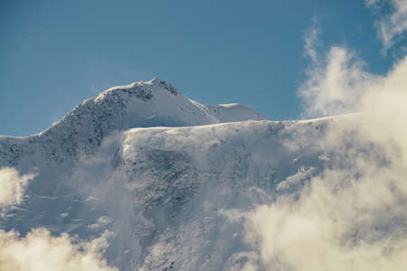 Minimalist view of snow-capped mountain wall in thick low clouds in sunshine. Scenic bright mountain landscape with white-snow peak among dense clouds in blue sky. Wonderful scenery with snowy top.の写真素材