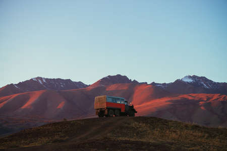 Mountain landscape with orange truck with view to pink magenta purple mountains with snow and sharp rocks on top under blue clear sky in sunset sunlight. Russia, Altai Republic, 20 September, 2020.のeditorial素材
