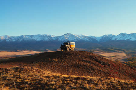 Truck on hill with view to great snow mountain range in sunset. Spectacular autumn landscape with truck on background of snowy mountains in red sunshine. Russia, Altai Republic, 20 September, 2020.のeditorial素材