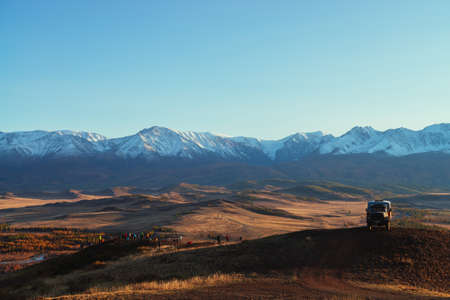 Truck and tourists on hill with view to red valley and great snow mountain range in red sunset sunshine. Spectacular sunny view to snowy mountains in autumn. Russia, Altai Republic, 20 September, 2020のeditorial素材