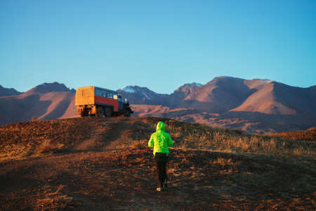 Orange truck and girl tourist in vivid green jacket on hill with view to great brown mountain with...