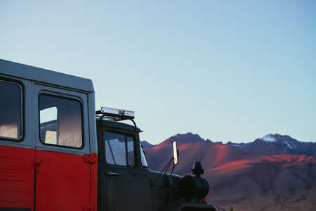Minimal closeup of orange truck with view to pink magenta purple mountains with snow and sharp rocks on top in blur under blue clear sky in sunset sunlight. Russia, Altai Republic, 20 September, 2020.のeditorial素材