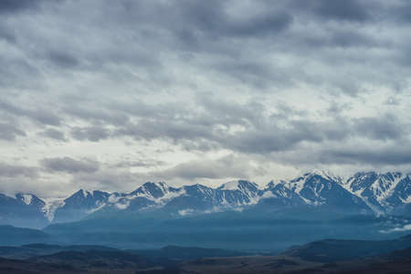 Scenic mountain landscape with great snowy mountain range among low clouds and green forest in valley at early morning. Atmospheric alpine scenery with blue white high mountain ridge under cloudy sky.の写真素材