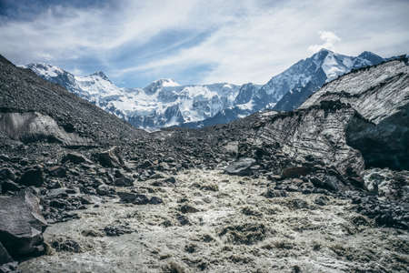 Scenic landscape with powerful mountain river beginning from glacier among large moraines on background of great snowy mountains. Beautiful scenery with glacier at source of turbulent mountain river.の写真素材