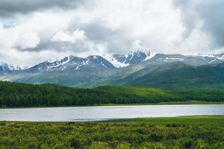 Dramatic landscape with mountain lake and forest on hills in sunlight and snowy mountains in low clouds in changeable weather. High mountains in cloudy sky and lake near green grasses and forest hillsの写真素材
