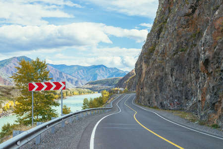 Vivid autumn landscape with mountain highway along big mountain river in sunshine. Bright alpine scenery with wide turquoise river and mountain road in autumn colors. Highway in mountains in fall timeの写真素材