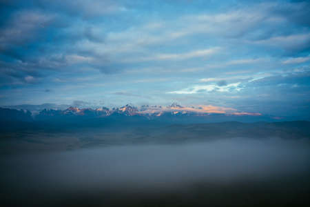 Scenic mountain landscape with great snowy mountain range lit by orange dawn sun above dense fog...