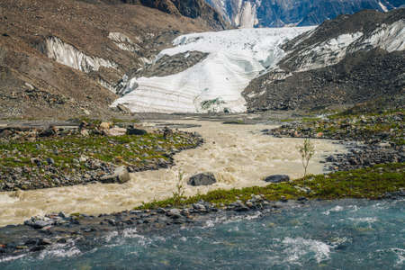Sunny alpine landscape with confluence of two various mountain rivers. Beautiful clear creek flows into dirty river. Colorful mountain scenery with confluence of two different rivers among rich flora.の写真素材