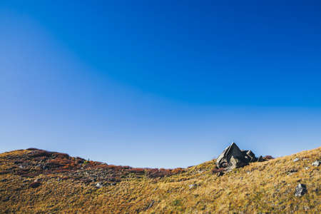 Minimal autumn landscape with pointy shattered stone on orange hill in sunlight under blue clear sky gradient. Colorful mountain scenery with hill in fall colors in sunshine. Autumn alpine minimalism.の写真素材