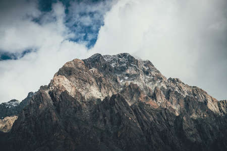 Mountain landscape with rocks with snow in sunlight and low clouds on top. Awesome rocky wall with sharp rocks in sunshine. Atmospheric mountain scenery with high rocky mountain pinnacle in clouds.の写真素材