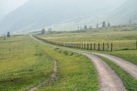 Beautiful green mountain landscape with long dirt road along fence and big mountains in fog. Atmospheric foggy mountain scenery with dirt road among hills and big mountains. Length road in countrysideの写真素材