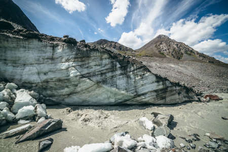 Scenic highlands landscape with mountain river beginning from glacier among large moraines on background of high mountain top. Beautiful scenery with glacier at source of mountain river and rock peak.の写真素材