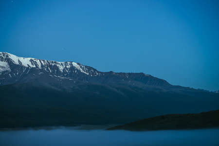 Atmospheric mountains landscape with dense fog and great snow mountain range under twilight sky. Alpine scenery with big snowy mountain ridge over thick fog in night. Snowy rocks above clouds in dusk.の写真素材